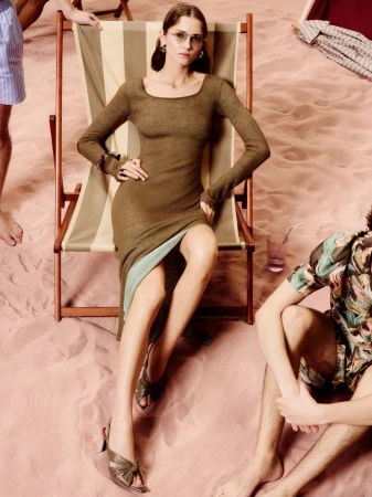 A woman in a brown dress sitting on a beach chair on a pink sand beach with two other partially visible people in the background.