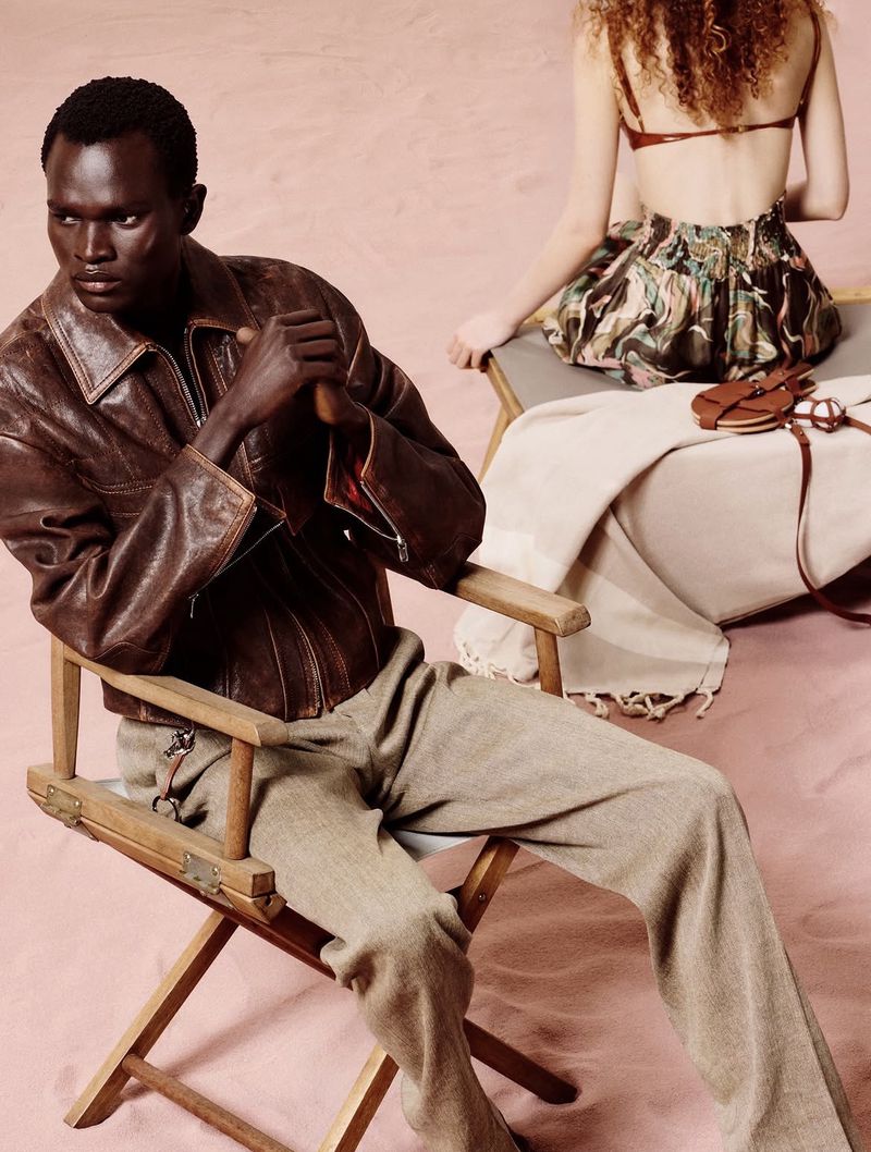 Fashion shot featuring an African male model in a brown leather jacket and tan trousers, seated on a director's chair, with another model.
