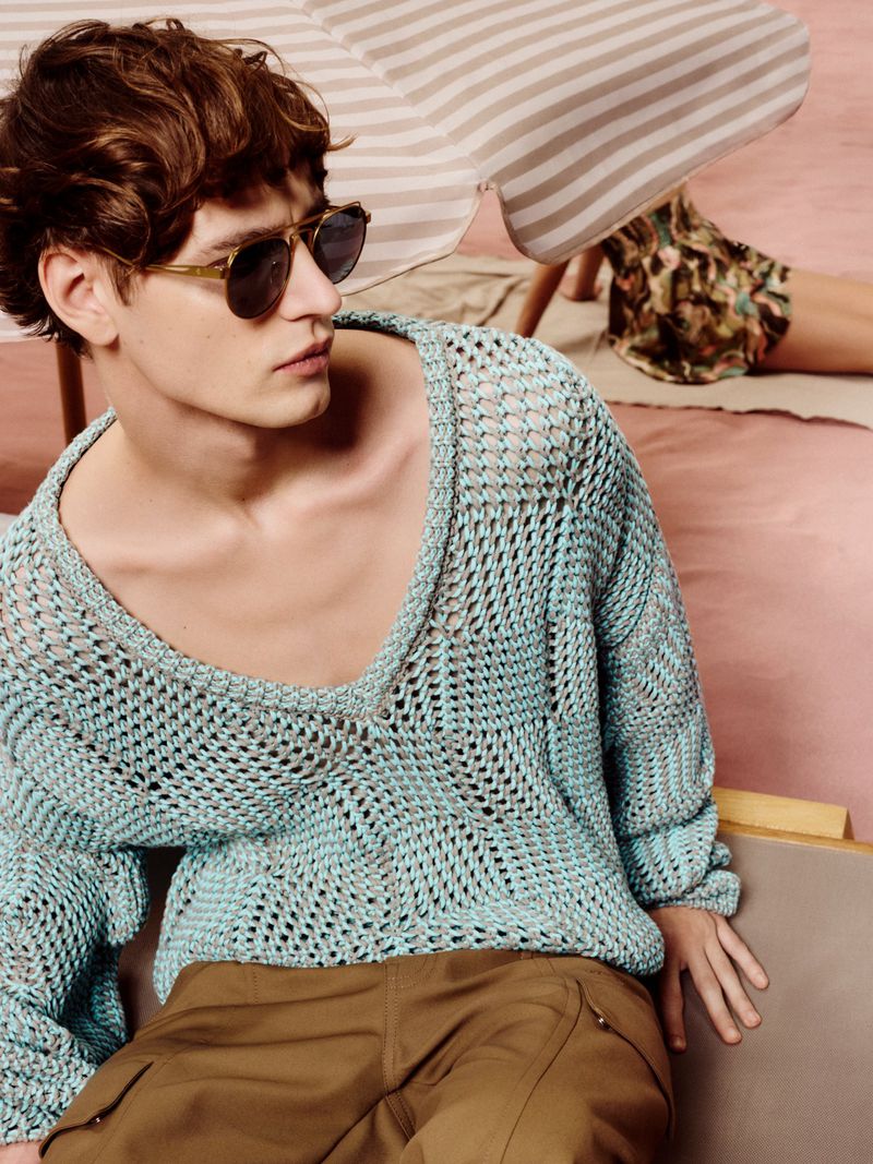 A young adult male models a light blue knit sweater and brown cargo pants in a bright, airy setting with a beach umbrella.