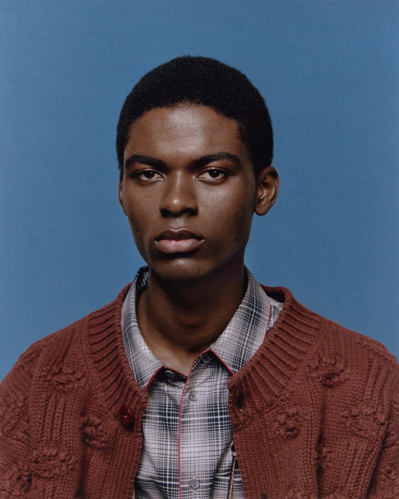 Portrait of an adult male with dark skin, wearing a plaid shirt and a brown knit cardigan, set against a solid blue background.