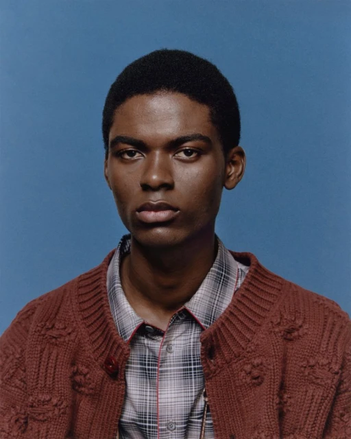 Portrait of an adult male with dark skin, wearing a plaid shirt and a brown knit cardigan, set against a solid blue background.