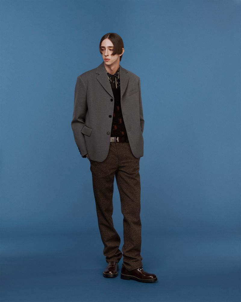 Full-length studio shot of a young adult male fashion model in a grey jacket and patterned brown pants against a blue backdrop.