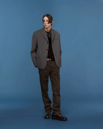Full-length studio shot of a young adult male fashion model in a grey jacket and patterned brown pants against a blue backdrop.