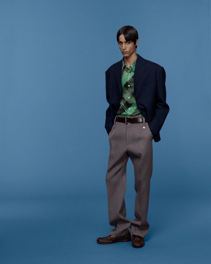 Full-length studio shot of a young adult male model wearing a green plaid shirt, gray pants, navy blazer, and leather loafers on blue backdrop.