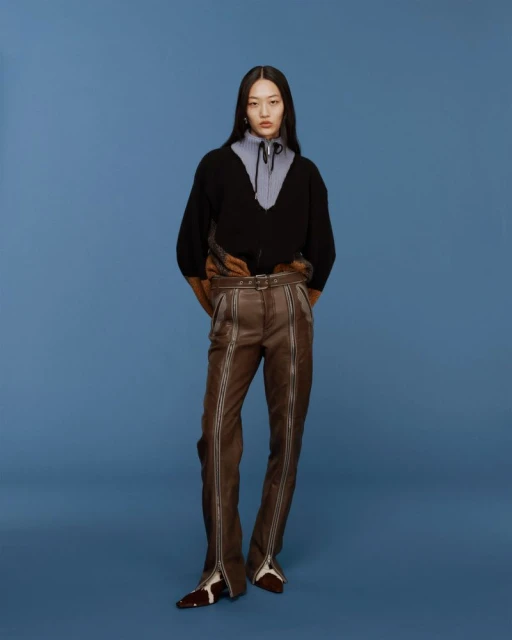 Full-length studio shot of an Asian female model wearing a black sweater, blue shirt, and brown pants with silver zippers on a blue backdrop.