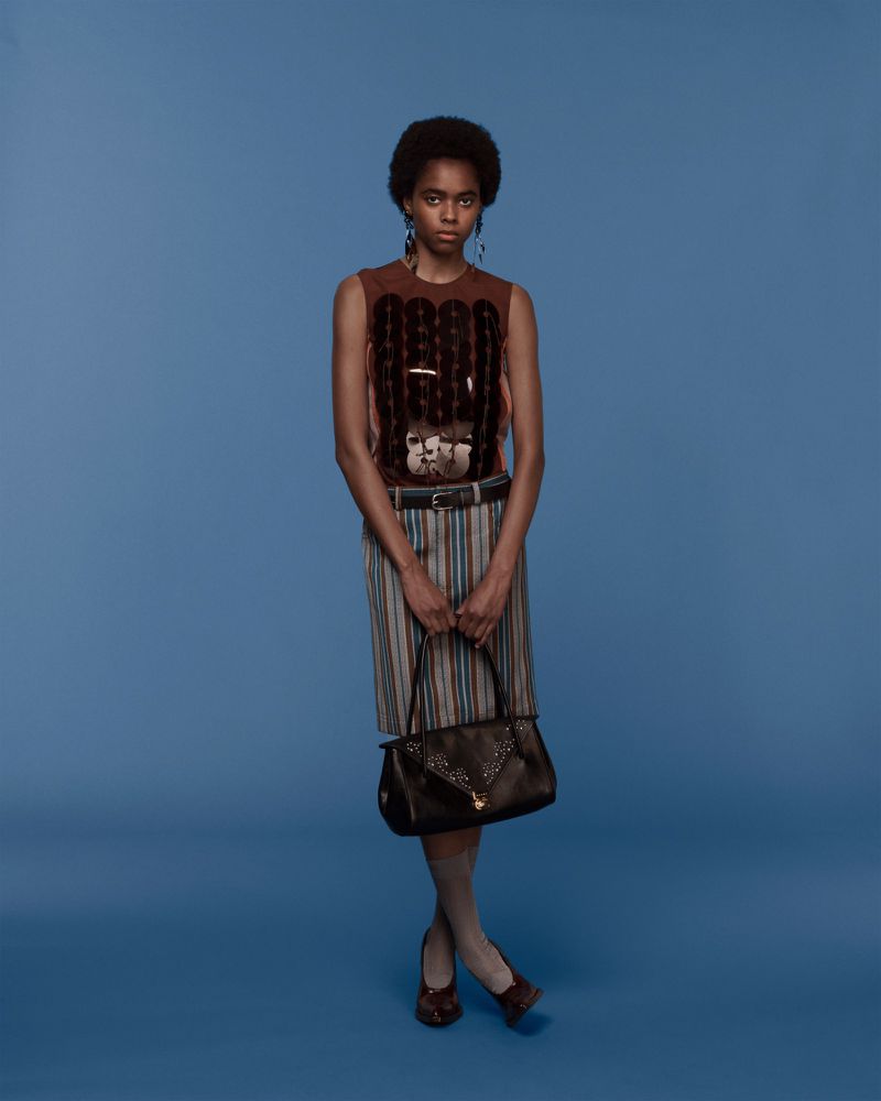Full-length shot of a Black woman in a patterned top and skirt, holding a brown purse, against a light-blue background in a studio setting.