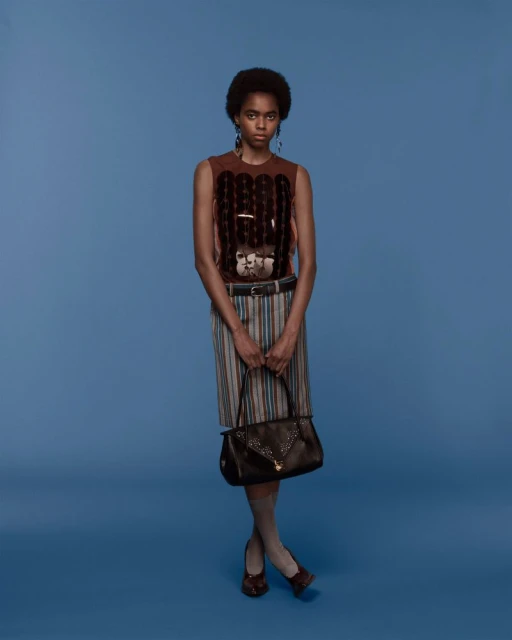 Full-length shot of a Black woman in a patterned top and skirt, holding a brown purse, against a light-blue background in a studio setting.