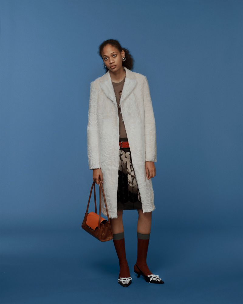 Studio shot of a female fashion model wearing a white overcoat, brown top, patterned skirt, brown purse, and brown socks against blue background.