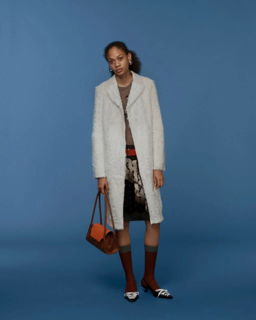 Studio shot of a female fashion model wearing a white overcoat, brown top, patterned skirt, brown purse, and brown socks against blue background.