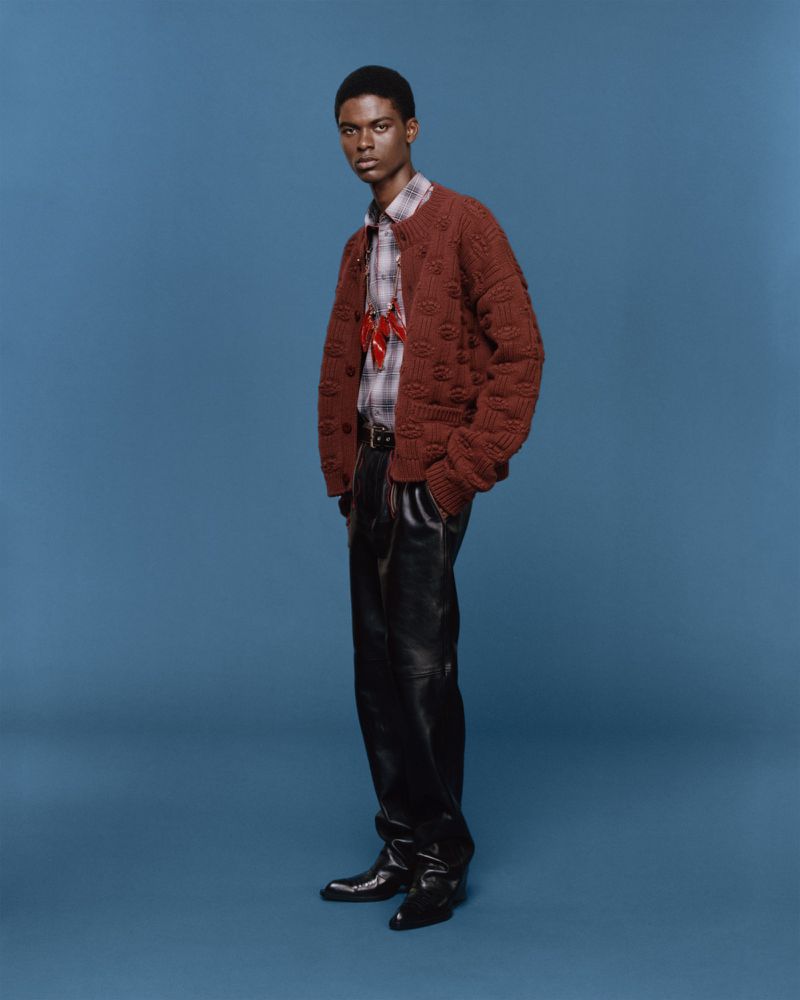 A full-length shot of a Black male model in a brown knit cardigan, plaid shirt, black leather pants and dress shoes, posing against a blue backdrop.