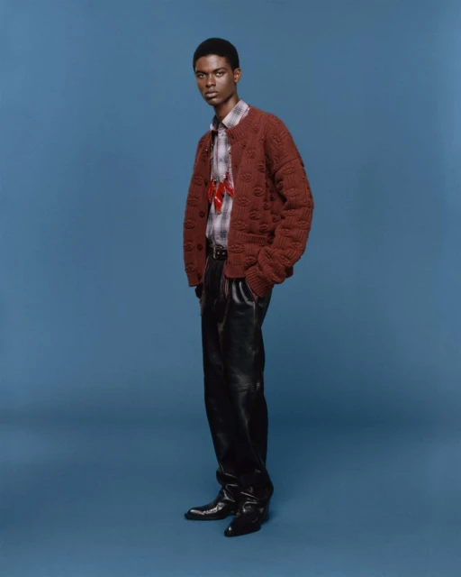 A full-length shot of a Black male model in a brown knit cardigan, plaid shirt, black leather pants and dress shoes, posing against a blue backdrop.