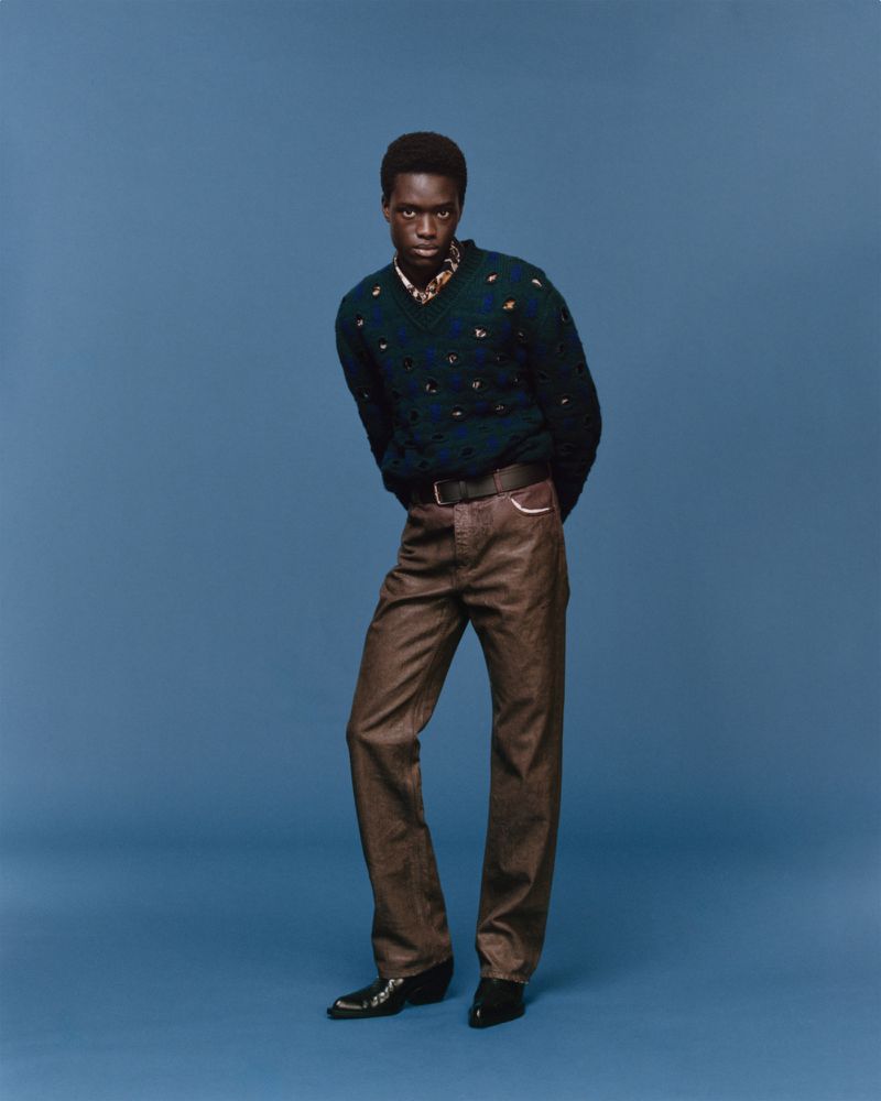 Full-length portrait of a Black male model in V-neck sweater and brown pants against a blue backdrop, showcasing fashion and style.