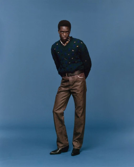 Full-length portrait of a Black male model in V-neck sweater and brown pants against a blue backdrop, showcasing fashion and style.