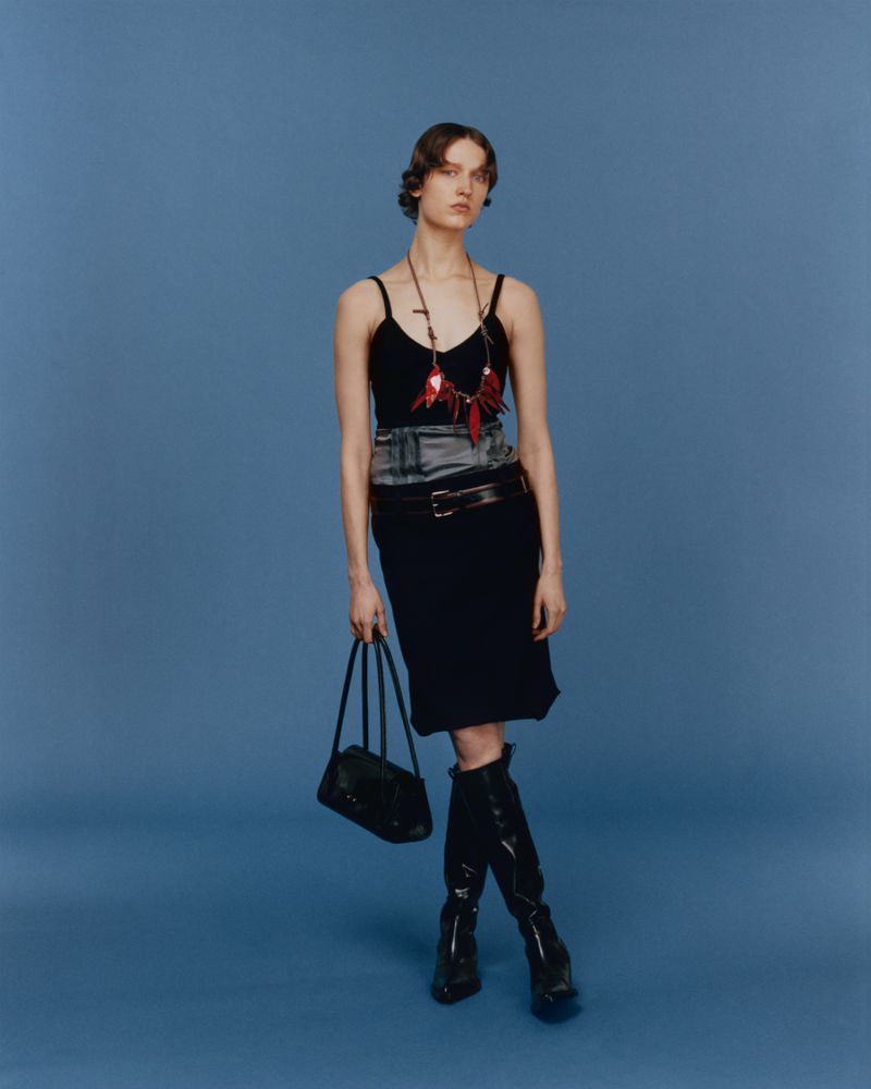 Fashion shot of a slender woman posing in a black top and skirt with tall black boots and a black purse on a blue backdrop.