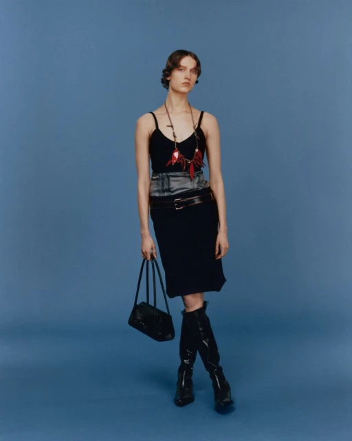 Fashion shot of a slender woman posing in a black top and skirt with tall black boots and a black purse on a blue backdrop.