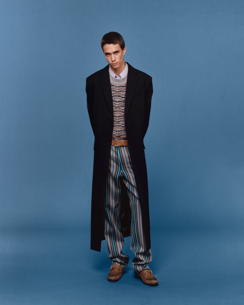 Full-length studio shot of a young adult man in a black coat, striped pants, and loafers against a solid blue background.