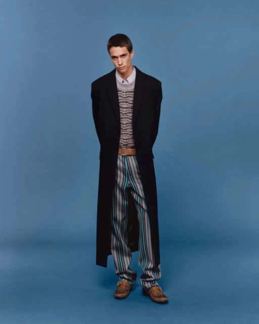 Full-length studio shot of a young adult man in a black coat, striped pants, and loafers against a solid blue background.