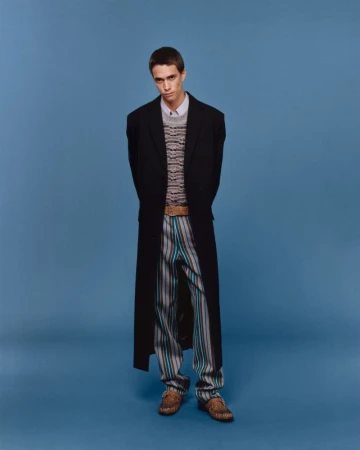 Full-length studio shot of a young adult man in a black coat, striped pants, and loafers against a solid blue background.