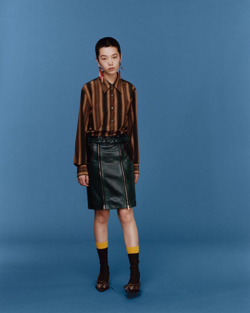 A woman with short dark hair wearing a striped shirt, dark skirt, socks, and shoes posing against a blue studio background.