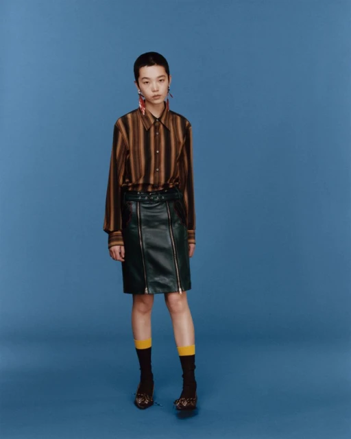 A woman with short dark hair wearing a striped shirt, dark skirt, socks, and shoes posing against a blue studio background.