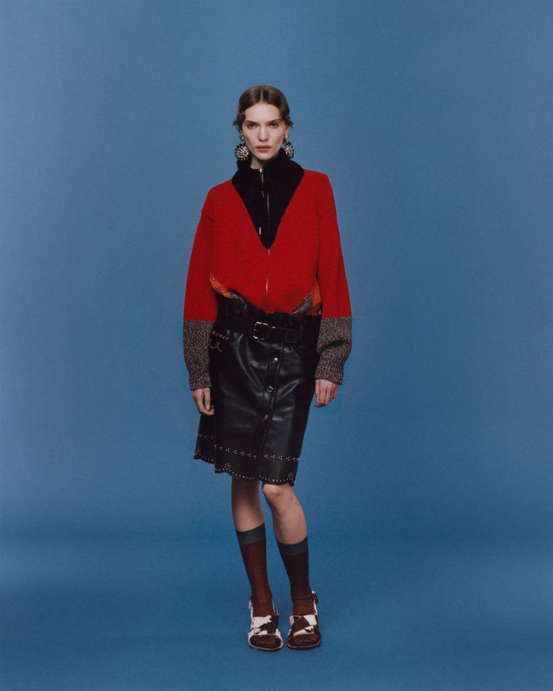 Fashion model posing in a red jacket, black leather skirt, socks, and shoes, set against a solid blue backdrop in a full studio shot.