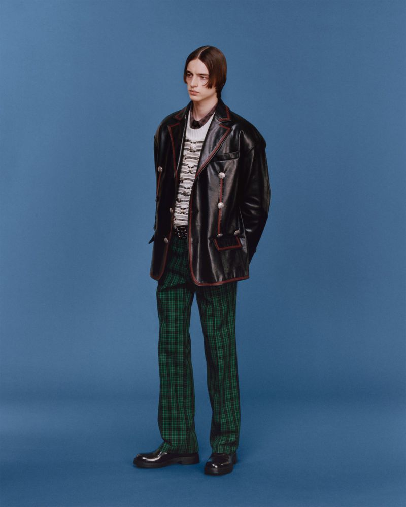 Full-length fashion portrait of a young adult male posing against a solid blue backdrop, wearing a black jacket and green plaid pants.