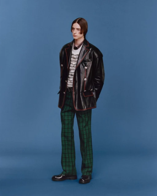 Full-length fashion portrait of a young adult male posing against a solid blue backdrop, wearing a black jacket and green plaid pants.