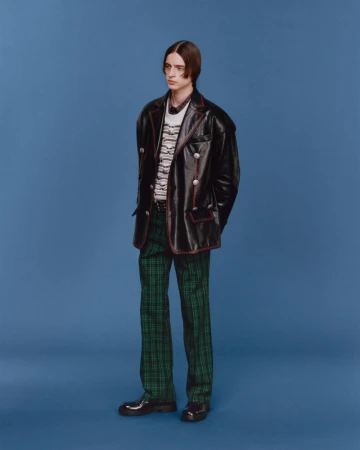 Full-length fashion portrait of a young adult male posing against a solid blue backdrop, wearing a black jacket and green plaid pants.