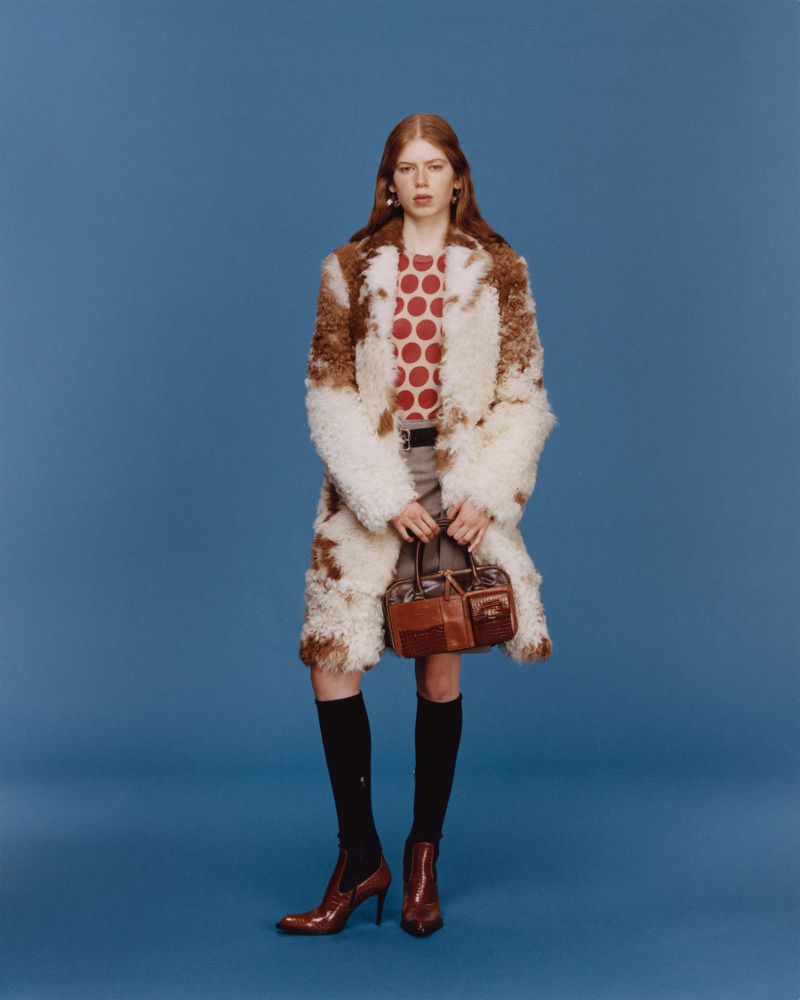 A stylish woman with red hair, wearing a fur coat and heeled boots, poses against a solid blue background, holding a satchel bag.