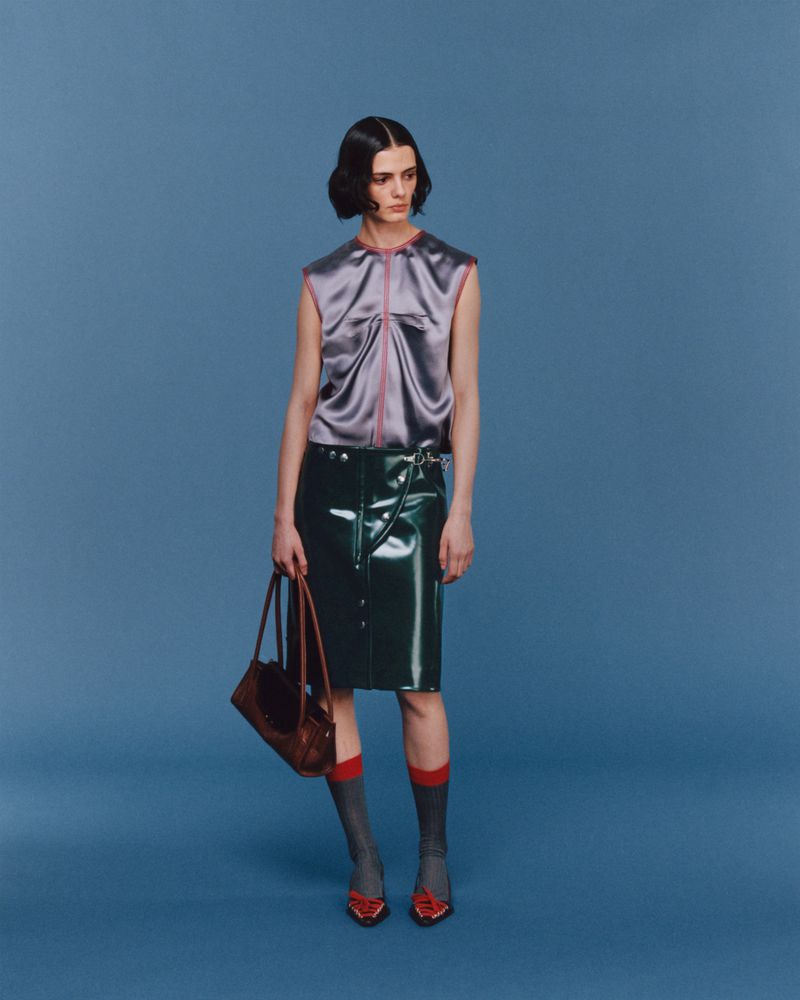 Fashion shot of a woman in a lavender sleeveless top, dark green skirt, patterned socks, and red shoes standing against a blue backdrop.