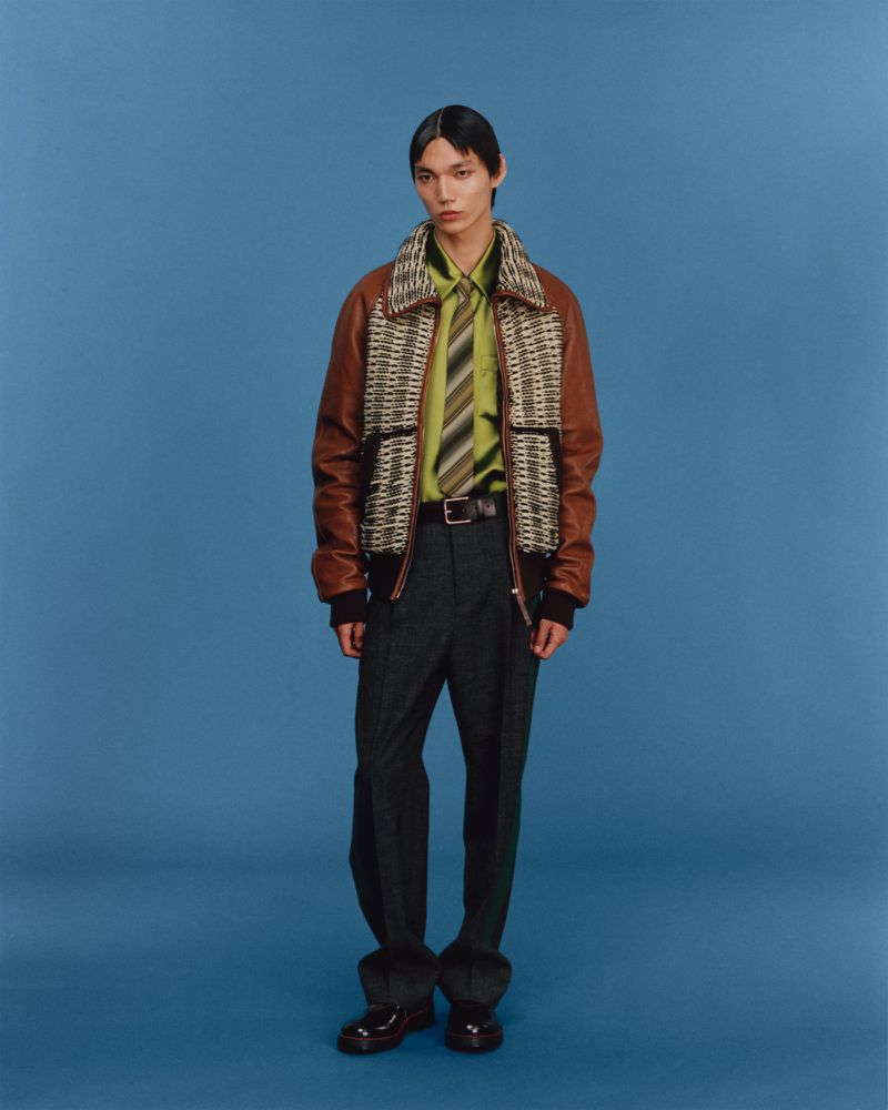 Full shot of a young adult male model wearing a brown leather jacket with a patterned insert, olive green shirt, striped tie, and dress pants.