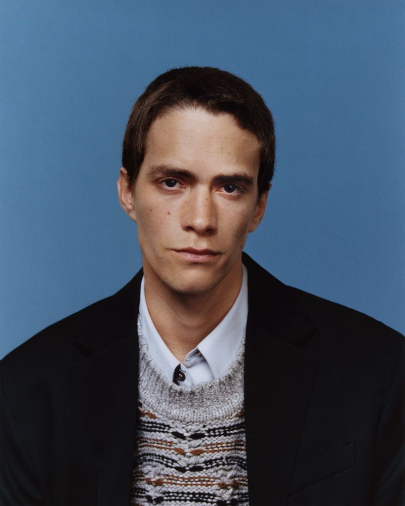 Portrait of a young adult man in a patterned sweater and blazer against a blue background, shot in a photo studio with soft, diffused lighting.