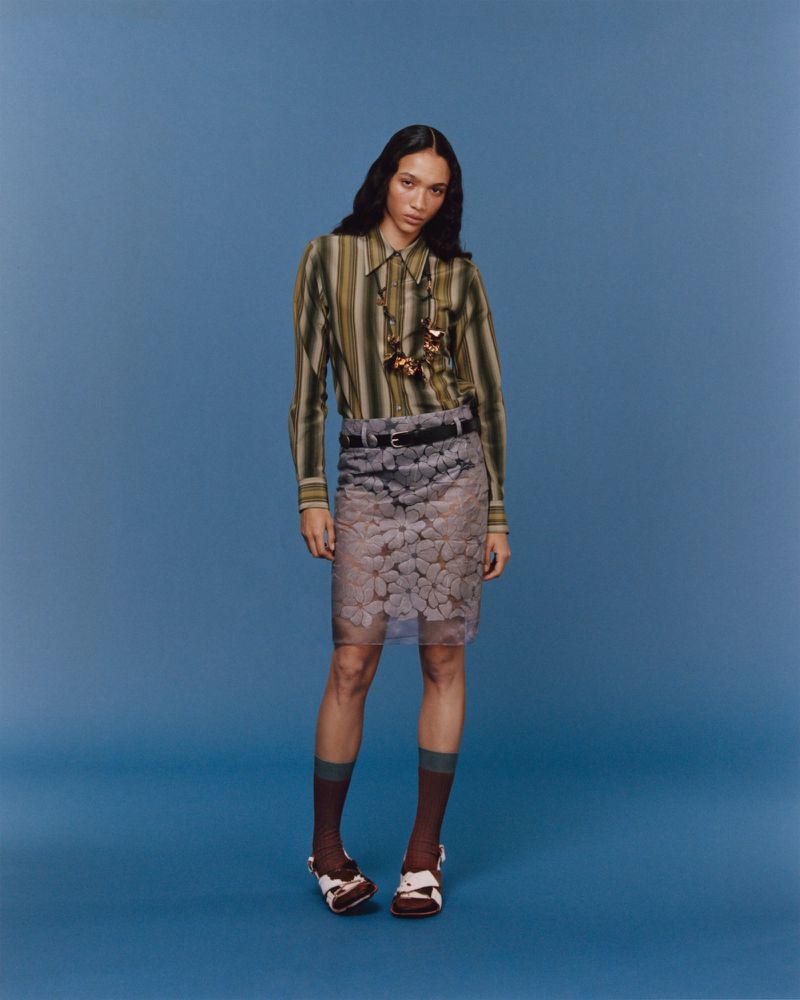 Full shot of an adult female fashion model with long dark hair wearing a striped button-up shirt and floral-patterned skirt against blue backdrop.