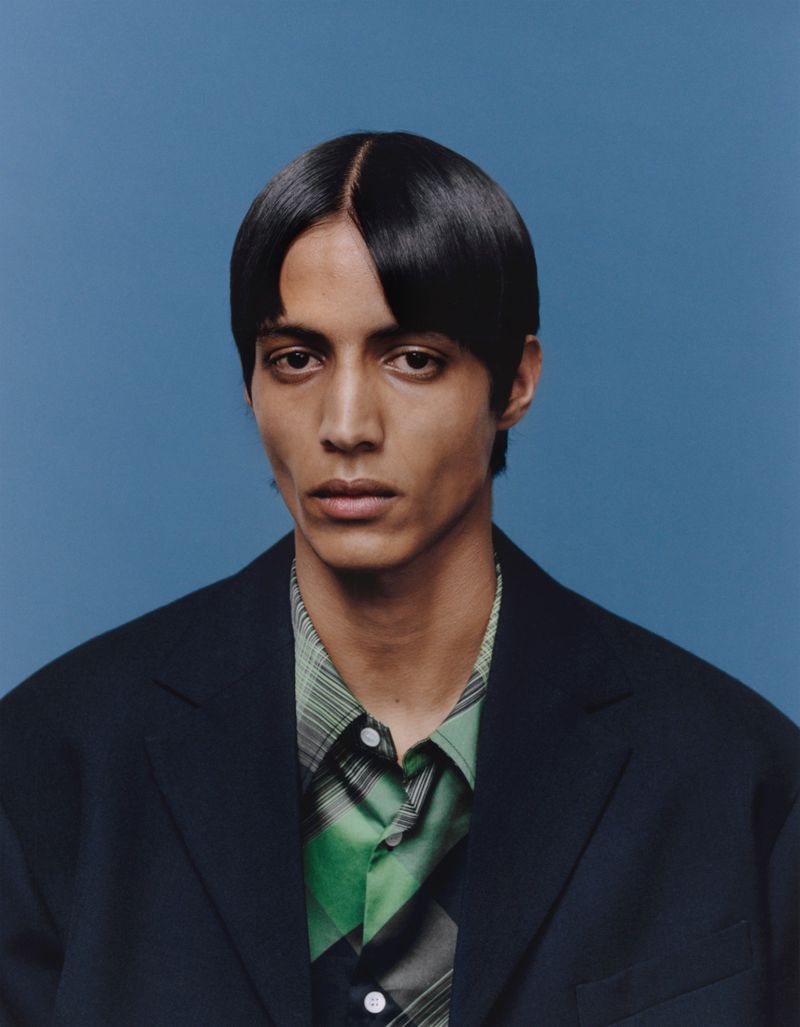 Portrait of a young adult male with dark hair, wearing a black jacket over a green and white plaid shirt, set against a solid blue background.