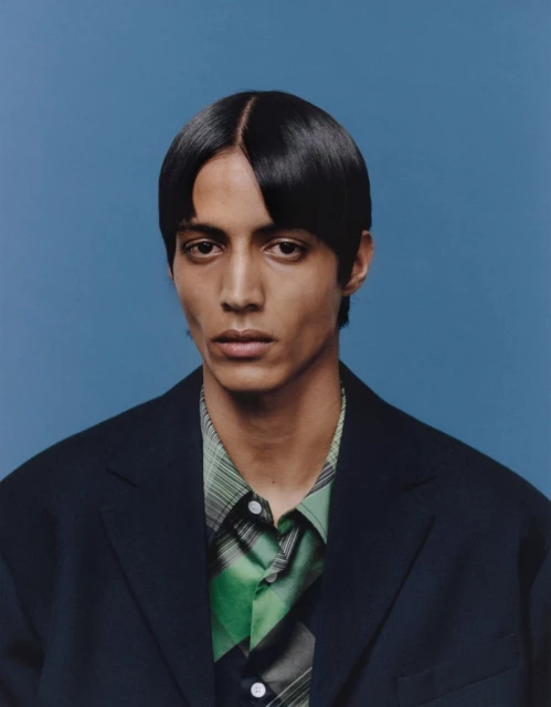 Portrait of a young adult male with dark hair, wearing a black jacket over a green and white plaid shirt, set against a solid blue background.