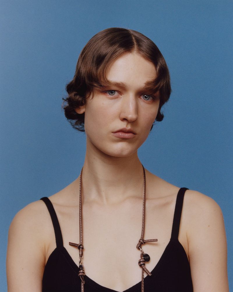 Portrait of a young woman with brown hair, wearing a black top and a rope necklace, against a solid blue background. Head and shoulders.