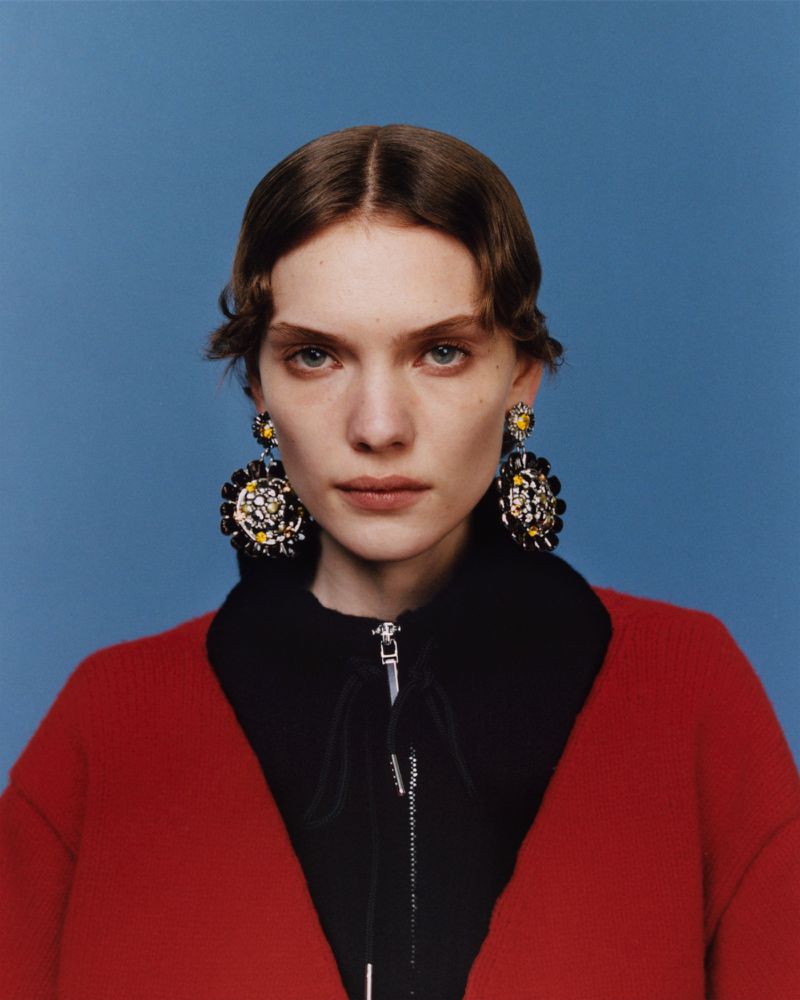 Close-up of a fair-skinned woman with reddish-brown hair wearing statement earrings, a red cardigan, and a black shirt against a blue backdrop.