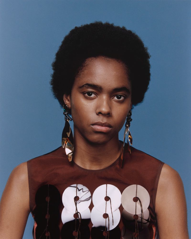 Headshot of a young Black woman with an afro, wearing a brown and white sequined top and long earrings against a light blue background.