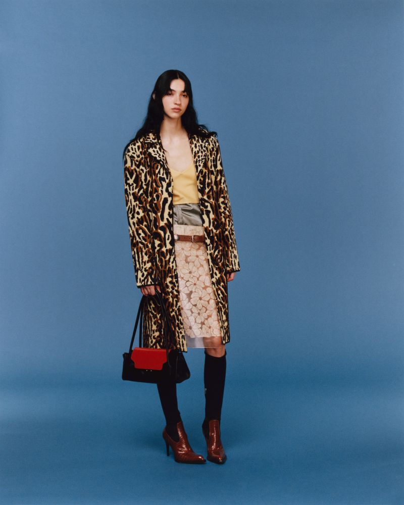 A woman in a leopard-print coat, skirt, boots, and a red and black bag stands in a studio against a solid blue backdrop.