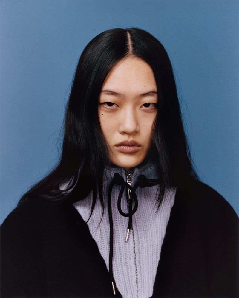 Headshot of an Asian woman with long black hair, wearing a black jacket and light purple sweater, set against a solid blue background.