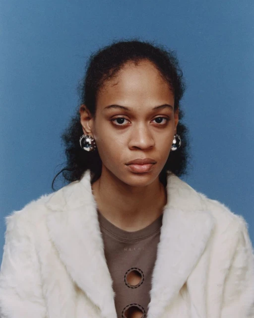 Portrait of a young woman with dark curly hair wearing a faux fur coat and large silver earrings against a blue background.