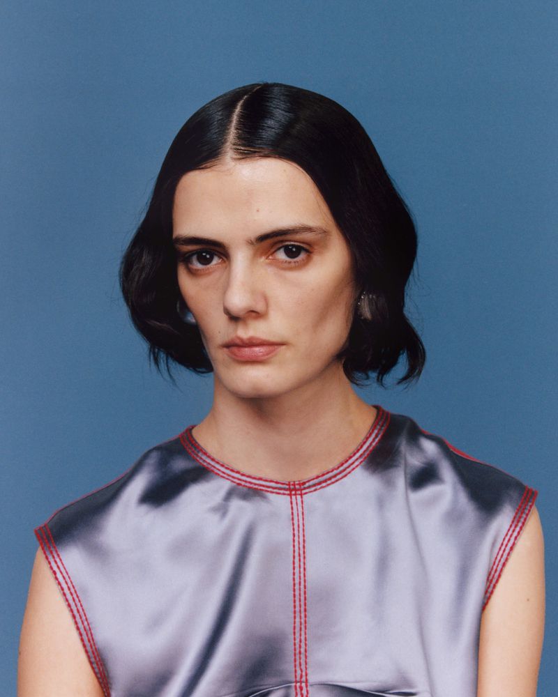 Close-up studio portrait of an adult female with fair skin, dark hair, wearing a sleeveless silver top with red stitching, against a blue background.