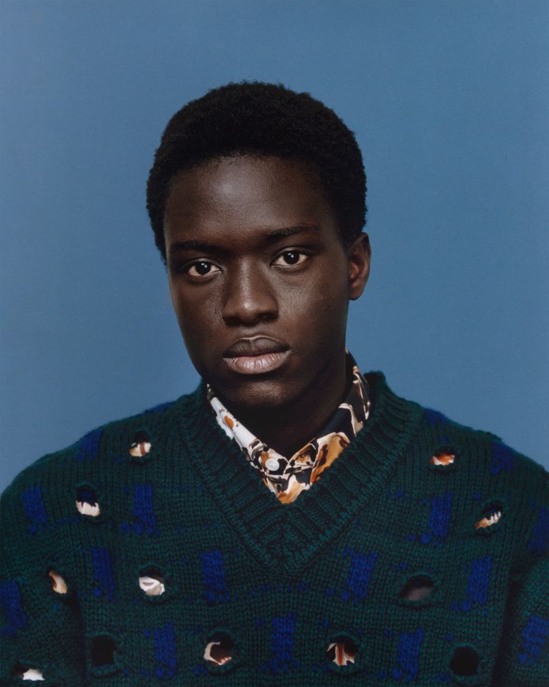 Portrait of a black male model wearing a green patterned sweater and collared shirt against a light blue backdrop in a photo studio.