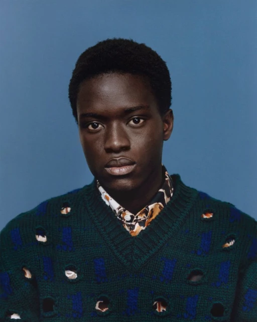 Portrait of a black male model wearing a green patterned sweater and collared shirt against a light blue backdrop in a photo studio.