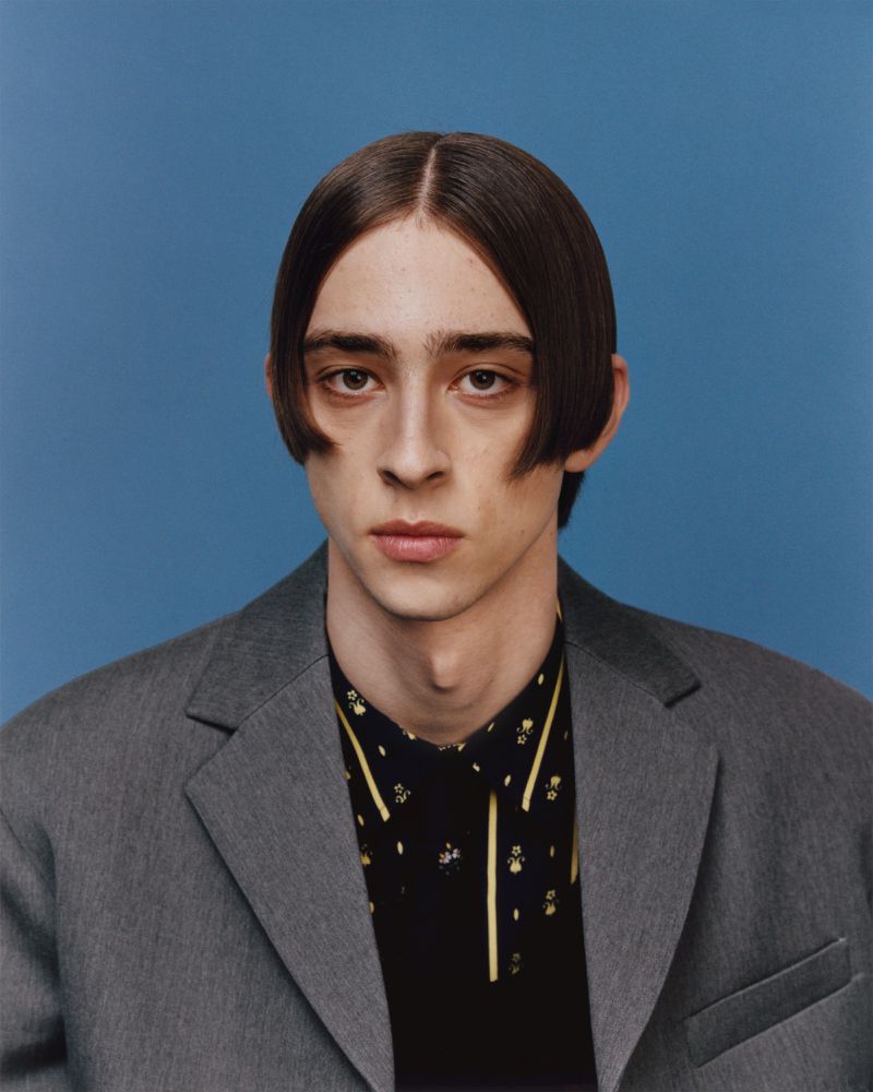 Close-up portrait of a young adult male with dark hair in a gray suit jacket and patterned shirt against a blue background.
