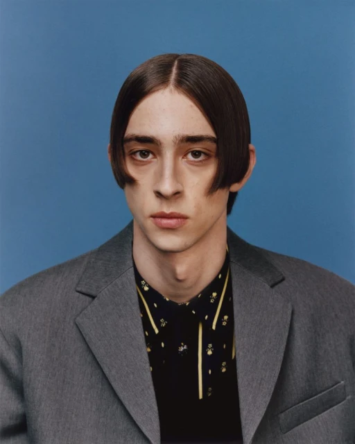 Close-up portrait of a young adult male with dark hair in a gray suit jacket and patterned shirt against a blue background.