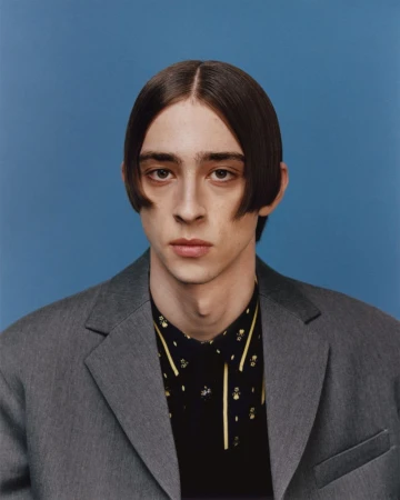 Close-up portrait of a young adult male with dark hair in a gray suit jacket and patterned shirt against a blue background.