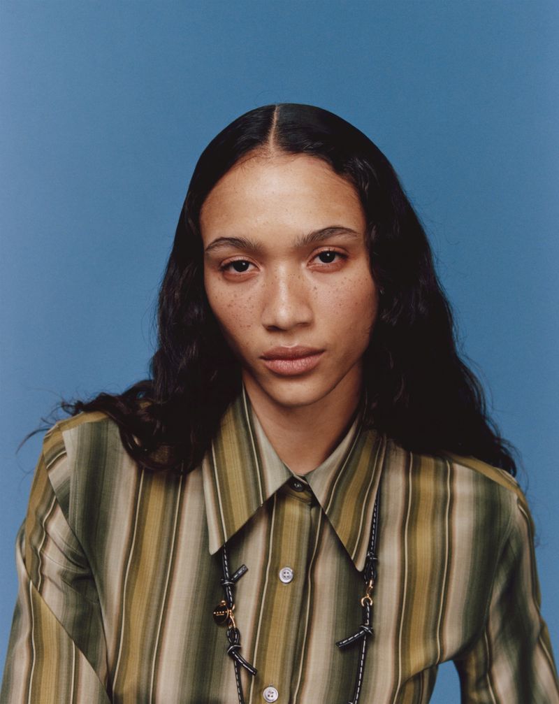 Portrait of a woman with dark wavy hair wearing a striped olive green and beige button-down shirt against a blue background.