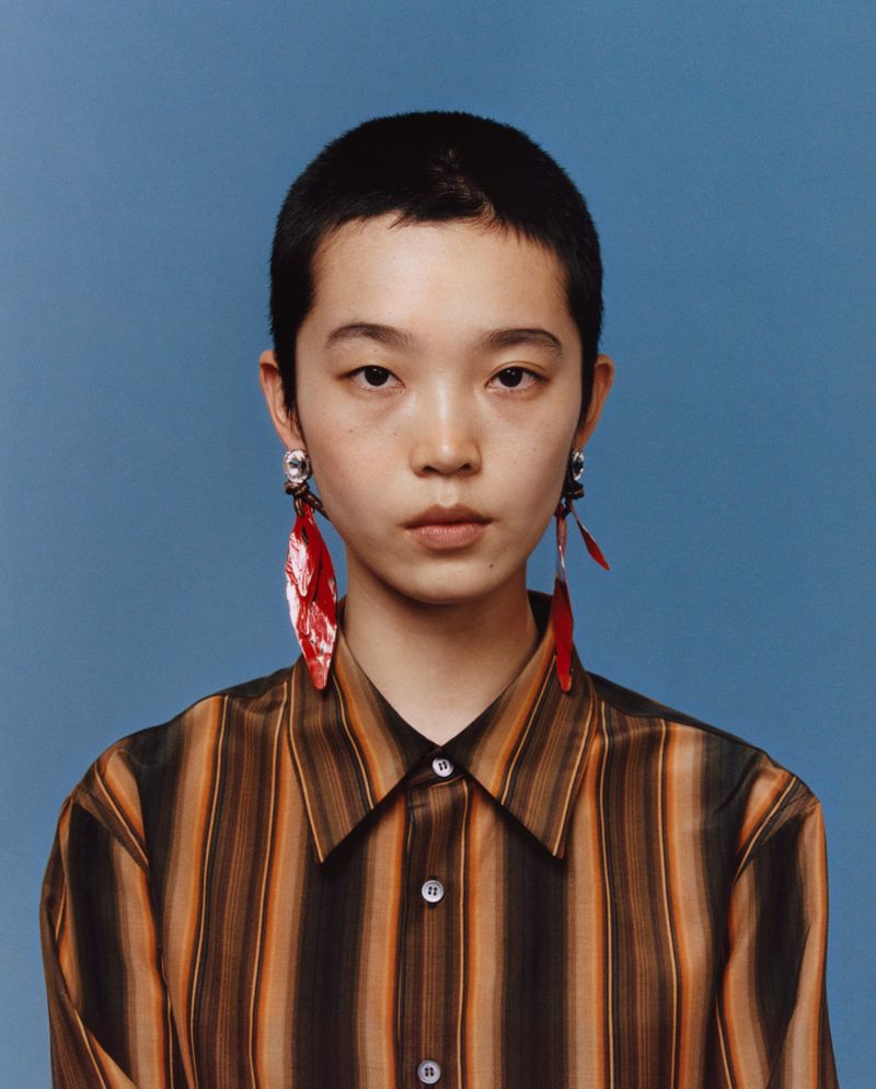 Portrait of a young adult with short hair wearing a striped shirt and statement earrings against a solid blue background.