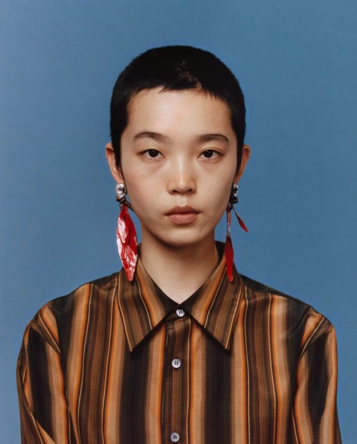 Portrait of a young adult with short hair wearing a striped shirt and statement earrings against a solid blue background.